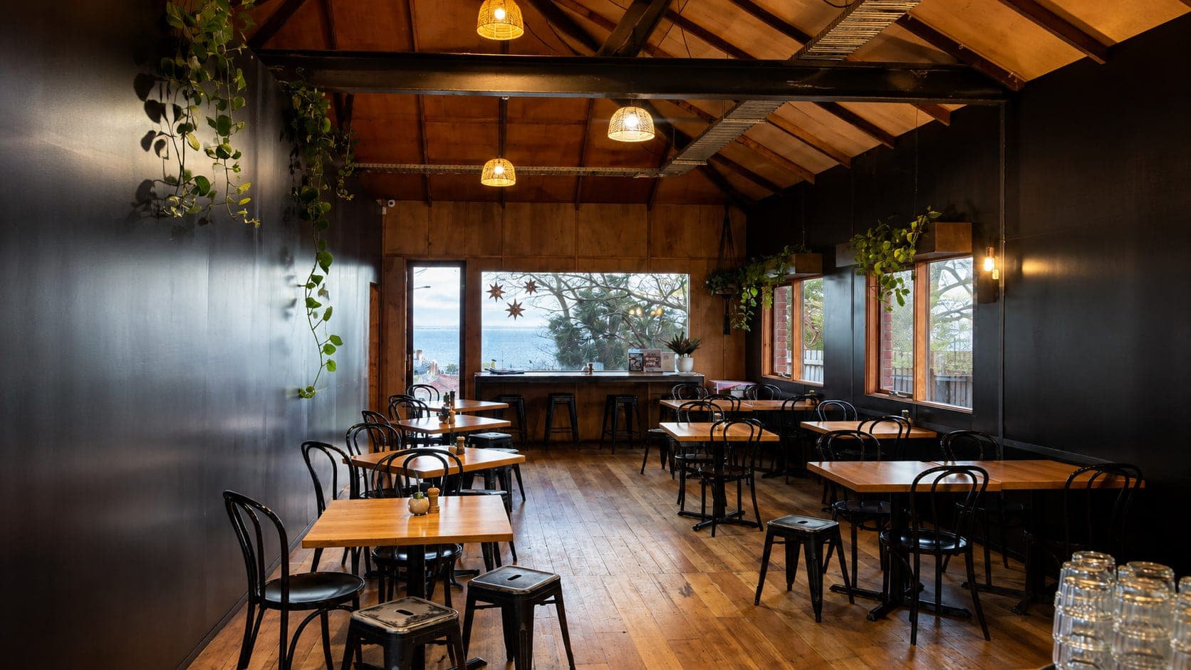The dining room's timber tables and bentwood chairs under rattan pendant lights, with Western Port Bay through the window
